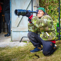 Ein sportlich gekleideter Mann mit auffälliger grüner Plüschjacke und grüner Mütze hockt auf einem Grasstück vor einem Schuppen und sieht konzentriert durch ein langes Zoom-Objektiv. Die Tür des Schuppens ist geöffnet, viel Gerümpel und zwei Personen befinden sich darin.