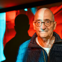 Ein kahlköpfiger älterer Herr in blauer Steppjacke steht vor einem bunten Bildschirm. Es gibt gleich zwei Schatten des Mannes auf dem Monitor.