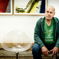 Ein älterer Mann sitzt in Jeans, grünem T-Shirt und Kapuzenjacke auf einem Stuhl in einem Atelier. Auf dem T-Shirt steht: "Life is good" Neben dem Mann steht ein transparenter, runder Loungestuhl. Im Hintergrund hängen zwei farbige Fotografien.