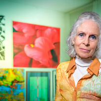 Hannelore Voltmer-Döbrich Portrait einer Frau mit weißen Haaren und orangefarbener Bluse: Sie steht vor einer Wand mit verschiedenfarbigen Gemälden und blickt in die Kamera.