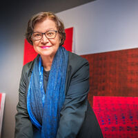 Eine ältere Dame mit Brille, dunklem Jackett und blauem Seidenschal steht ein wenig vorgebeugt in einem Ausstellungsraum und schaut genau in die Kamera. An der Wand und auf dem Boden sind etliche große, rote und rotblaue abstrakte Gemälde zu sehen. Im Hintergrund ein Sprossenfenster und ein Stuhl.