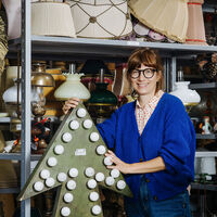 Eine Frau steht vor einem großen Lagerregal, das eng mit den unterschiedlichsten Lampen und Lampenschirmen bepackt ist. Mit den Händen stützt sie sich auf einen bronzefarbenen Pfeil, der mit Glühlampen umrandet ist.