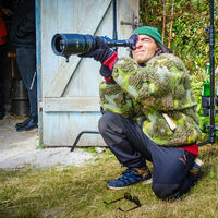 Fatih Akin Ein sportlich gekleideter Mann mit auffälliger grüner Plüschjacke und grüner Mütze hockt auf einem Grasstück vor einem Schuppen und sieht konzentriert durch ein langes Zoom-Objektiv. Die Tür des Schuppens ist geöffnet, viel Gerümpel und zwei Personen befinden sich darin.