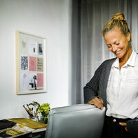 Sabrina Müller-Wüsthoff Eine Frau in weißer Bluse und grober Strickjacke neigt schmunzelnd den Kopf nach unten, die Augen sind geschlossen. Sie lehnt an einen Schreibtischstuhl. Auf dem Tisch verschiedene Pinsel und Stifte sowie ein aufgeschlagenes Buch mit Aquarellen. An der Wand zwei gerahmte Aquarelle. Eines zeigt zwei Wellensittiche.