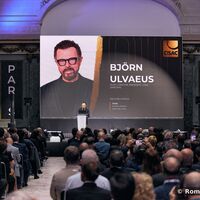 Der CISAC-Präsident Björn Ulvaeus hält die Eröffnungsrede zur Generalversammlung anlässlich des 100. Geburtstags von CISAC in Paris. Auf einer großen Leinwand ist das Foto und der Name des Redners groß eingeblendet. Davor steht Björn Ulvaeus an einen Rednerpult. Der prunkvolle Kuppelsaal ist gut gefüllt. 