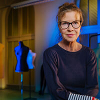 In einem Saal mit hohen Decken und großen Fenstern stehen mehrere Kleiderpuppen über den Raum verteilt, die farbig angestrahlt werden. Sie tragen kein Kostüm. Im Vordergrund steht eine Frau und schaut direkt in die Kamera.