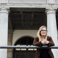 Eine Frau mit langen blonden Haaren lehnt sich über die Eisenbrüstung eines klassizistischen Gebäudes und schaut in die Kamera, flankiert von zwei griechischen Säulen. Im Hintergrund schwere, halbrunde Eichentüren, Kassettendecke und zwei Laternen.