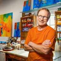 Jörgen Habedank Ein schalkhaft grinsender Herr mit Brille im auffällig orangefarbenen T-Shirt steht in einem Maleratelier mit bunten Bildern, nach Farbe sortierten Ölfarben und Pinseln.
