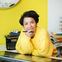 Portrait einer Frau im gelben Stickpullover, die sich auf ein Grafiktablett stützt und direkt in die Kamera sieht. Im Hintergrund eine alte Schreibmaschine und Bücher.