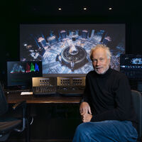 Ein Mann sitzt in einem abgedunkelten Raum vor einem Bedienpult für Filmbearbeitung, der Kamera zugewandt. Auf einem großen Monitor ist ein Filmausschnitt zu sehen. Der Ausschnitt zeigt ein großes Oktagon mit einem runden Aufbau in der Mitte, der von Engeln bewacht wird, darauf ein Harfenist.