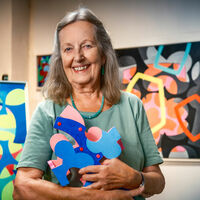 Barbara Jäger Eine ältere Frau mit langem grauen Haar hält ein buntes, florales dreidimensionales Puzzelteil aus Holz im Arm. Auf einer Staffelei und an den Wänden hängen verschiedene bunte Kunstwerke, die ähnliche Muster zeigen.