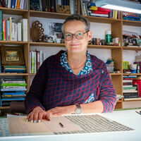 Die Künstlerin sitzt an ihrem Schreibtisch und sieht direkt in die Kamera. Vor ihr liegen Vorlagen mit handgeschriebenen Buchstaben, darauf Millimeterpapier und ein Bleistift. Hinter ihr steht ein großes Regal mit Büchern und Farben.