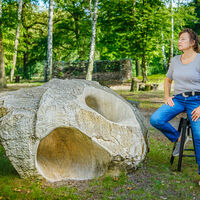 Birgit Cauer In einem Laubwald sitzt eine Frau auf einem Drehhocker vor einer eiförmigen Steinskulptur.