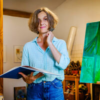 Die Künstlerin in Jeans und blauer Bluse steht in ihrem Atelier neben einer Holzsäule und hält einen aufgeschlagenen Katalog in der Hand. Auf einer Staffelei steht ein grünes Gemälde, das Pflanzenteile zeigt. An den Wänden hängen weitere Bilder, auf Tischen stehen Pinsel, Stifte und Farben.