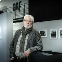 Rolf Nobel Ein älterer Herr mit weißen Haaren und weißem Bart, Brille, kariertem Hemd, Jacke und Schal steht in einer Fotoausstellung an ein Desk gelehnt. Eine Galerie mit Geländer ist im Hintergrund zu sehen. An der oberen Wand steht die Abkürzung "GAF". Das Bild ist in schwarz-weiß gehalten mit wenig Farbe.