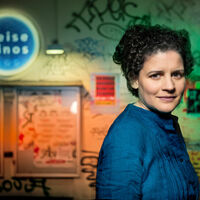 Junge Frau mit dunklem Lockenschopf und blauer Bluse steht vor der Hauswand eines Kinos. Auf einer Leuchtreklame steht "Zeise Kinos", darunter zwei Filmplakate. Die Wand ist mit Graffiti besprüht.