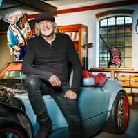 In einer Garage sitzt ein älterer Mann mit halblangem, blonden Haar, lässiger Kleidung und Schiebermütze entspannt auf dem Kotflügel eines eleganten Sportwagens mit offener Motorhaube. Im Hintergrund die Comicfigur „Werner“ mit Bierflasche als Pappfigur und eine E-Gitarre mit der Aufschrift "Bölkstoff". Ein aufgesprühtes Peace-Symbol mit einem Herzen mit Fischflossen. 