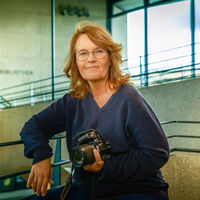 Eine Frau mit langen Haaren und Brille sitzt in einem Treppenhaus aus Sichtbeton, mit Metallgeländer und Fensterfront. Sie hält eine Kamera von Nikon in der Hand. Auf einer Wand im Hintergrund ist undeutlich das Wort "Bibliothek" zu lesen.