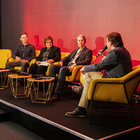 Auf einem Podium sitzen drei Männer und eine Frau in gelben Loungesesseln in eine Diskussion vertieft. Der Mann ganz rechts spricht in ein Mikrophon; die anderen drei hören ihm aufmerksam zu.