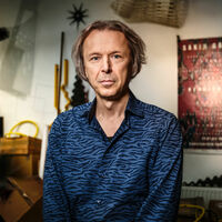 Şakir Gökçebağ Portrait eines Mannes im blau gemusterten Hemd. Verschiedene Gegenstände stehen und hängen an der Wand: eine Leiter, ein Plakat, ein Korb, einige Kisten.