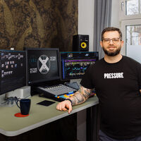 Ein junger Mann in schwarzem T-Shirt mit dem weißen Aufdruck "Pressure" steht an einem Stehpult, den tätowierten Arm lässig aufgestützt und sieht lächelnd in die Kamera. Auf dem Tisch stehen drei Bildschirme sowie ein Mischpult. Auf dem mittleren Bildschirm ist ein weißes Kreuz in einem Kreis abgebildet sowie der zweimalige Schriftzug "Picture Start". Auf zwei gesonderten Ständern sind zwei Lautsprecher angebracht. 