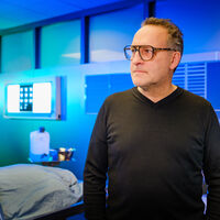 Ein Mann mit Brille schaut zur Seite. Er steht in einem Sektionssaal der Rechtsmedizin, der in ein fluoreszierendes blaues Licht getaucht ist. Man sieht eine angedeutete zugedeckte Leiche, ein Mikroskop, ein paar Blutproben, ein Kanister mit Desinfektionsmittel, eine OP-Lampe, medizinische Instrumente. 