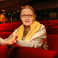 Eine ältere Dame mit Brille, zurückgebundenen langen Haaren und um die Schultern gelegtem Schal sitzt in einem Kinosaal mit roten Sitzen. Sie redet lebhaft mit den Händen und schaut direkt in die Kamera.