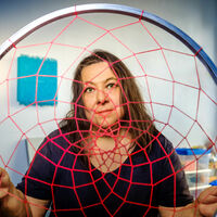 Irene Anton Eine Frau mit langen Haaren schaut durch ein großes, rotes Traumfänger-Netz mit einem Edelstahlrahmen in die Kamera. Im Hintergrund sind weitere textile Kunstwerke in bunten Farben und einiges Material in Kisten zu sehen.