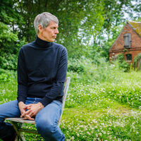 Ein schlanker Mann mit grauen Haaren, in Jeans und blauem Rollkragenpullover sitzt auf einem Gartenstuhl auf einer grünen Wiese. Im Hintergrund ist ein rotes Backsteinhaus sowie viele Laubbäume zu sehen. Der Mann schaut der Kamera abgewandt zur Seite.