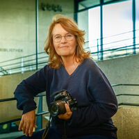 Caroline Link Eine Frau mit langen Haaren und Brille sitzt in einem Treppenhaus aus Sichtbeton, mit Metallgeländer und Fensterfront. Sie hält eine Kamera von Nikon in der Hand. Auf einer Wand im Hintergrund ist undeutlich das Wort "Bibliothek" zu lesen.