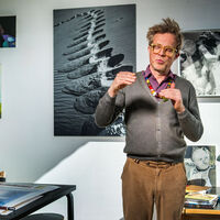 Mann mit gelber Brille, bunter Perlenkette und strubbeligem Haar redet lebhaft mit den Händen, die Augen geschlossen. An der Wand hängen verschiedene Schwarz-Weiß-Fotografien und farbige Gemälde. Auf einem Besprechungstisch liegen Aquarelle und eine Pfeife mit Tabak. 