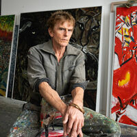 Torsten Schlüter Ein Mann sitzt in einem Atelier auf einem umgedrehten Stuhl, die Hände überkreuz und blickt in die Kamera. Seine Hose ist mit bunter Farbe bekleckst. Drei großformatige abstrakte Ölgemälde stehen hinter ihm an der Wand. Auf einem Bild ist ein Tigerkopf zu sehen.