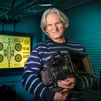Ein älterer Mann mit längerem grauen Haar hält eine Fernsehkamera im Arm. In dem abgedunkelten Technikraum hängt eine leuchtende Focus chart zur Kameraeinstellung.