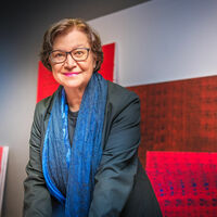 Miro Zahra Eine ältere Dame mit Brille, dunklem Jackett und blauem Seidenschal steht ein wenig vorgebeugt in einem Ausstellungsraum und schaut genau in die Kamera. An der Wand und auf dem Boden sind etliche große, rote und rotblaue abstrakte Gemälde zu sehen. Im Hintergrund ein Sprossenfenster und ein Stuhl.