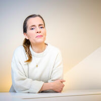 Anne-Ulrike Thursch Eine Frau mit Zopf und weißem Pullover steht vor einem cremefarbenem Hintergrund und lehnt sich auf eine weiße Brüstung. Den Kopf im Nacken schaut sie direkt in die Kamera. Auf der Brüstung steht ein Glaskasten mit einem Buddha-Kopf und einer Kerze. Der ganze Raum ist sehr hell.