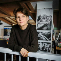 Sabine von Bassewitz Eine Frau stützt sich auf ein Treppengeländer in einem Dachgeschoss, das als Büro genutzt wird, und sieht verträumt zur Seite. An einem Pfeiler hängen Schwarz-Weiß-Fotografien. Ein Schreibtisch mit großem Bildschirm steht im Hintergrund.