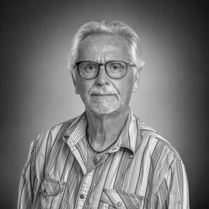 Portrait eines weißhaarigen Herrn mit Brille und Bart im gestreiften Hemd und Kette mit auffälligem Stein. 