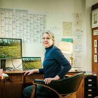 Nana Petzet Eine Frau sitzt an einem Schreibtisch und hat sich zur Kamera umgedreht. Auf zwei Bildschirmen ist jeweils eine grüne Wiese zu sehen. Neben dem Schreibtisch steht ein gutgefülltes Bücherregal und ein Globus.