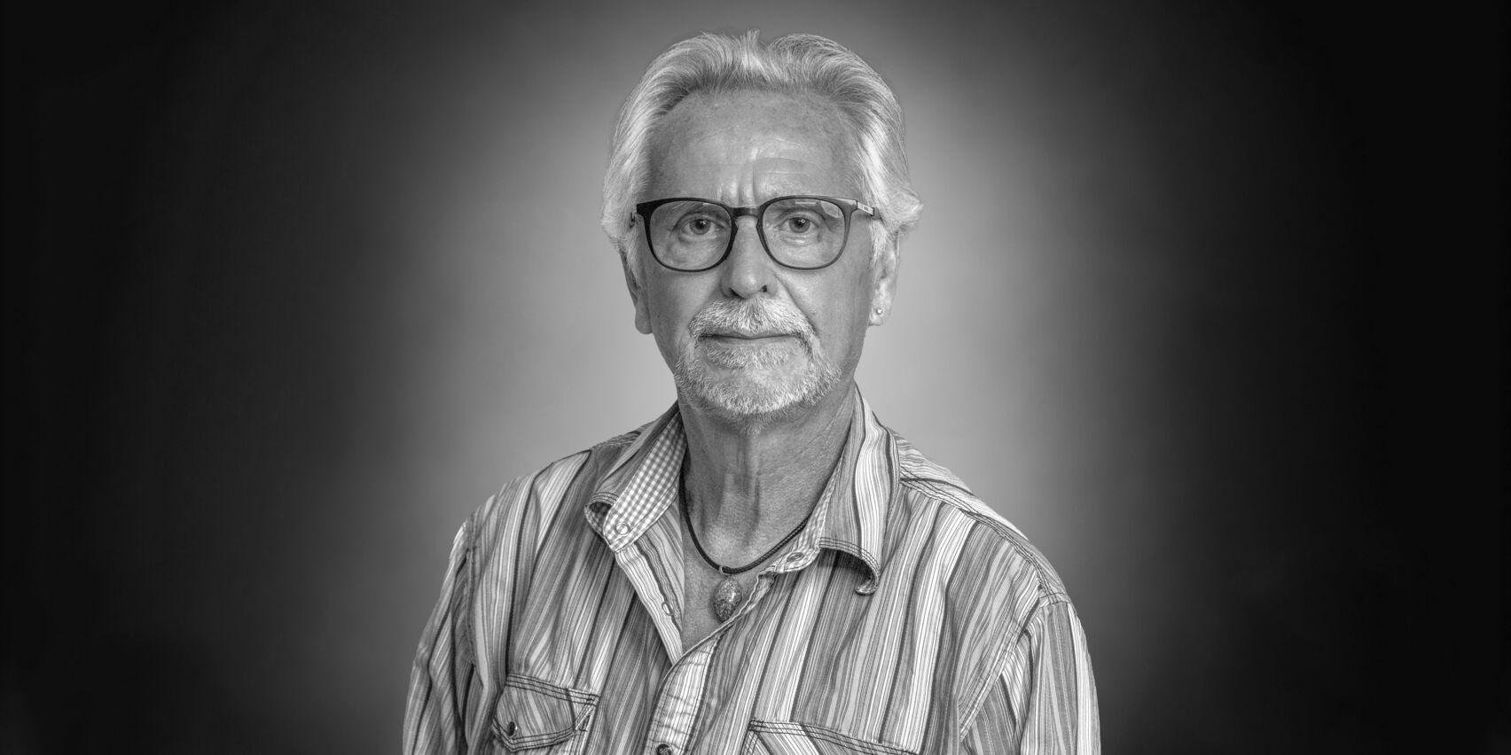 Portrait eines weißhaarigen Herrn mit Brille und Bart im gestreiften Hemd und Kette mit auffälligem Stein.