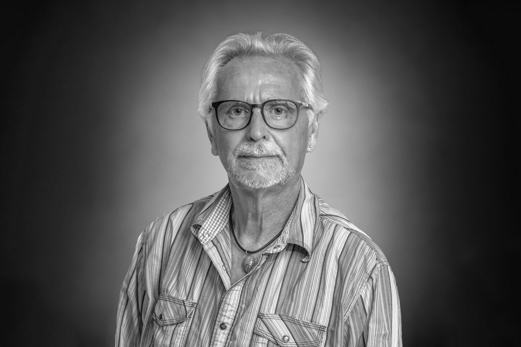 Portrait eines weißhaarigen Herrn mit Brille und Bart im gestreiften Hemd und Kette mit auffälligem Stein.