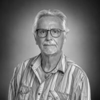 Portrait eines weißhaarigen Herrn mit Brille und Bart im gestreiften Hemd und Kette mit auffälligem Stein. 