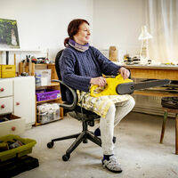 Cornelia Konrads Eine Frau sitzt auf einem Schreibtischstuhl in der Mitte eines Ateliers. Sie hält eine gelbe Kettensäge auf dem Schoß. In verschiedenen Kisten und Kleinmöbeln wird Werkzeug aufbewahrt. Ein Arbeitstisch ist mit Spanplatten und anderem Material belegt.