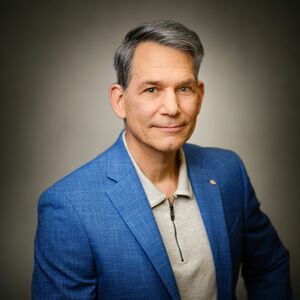 Portrait eines grauhaarigen Herrn in hellblauem Jackett und beigefarbenem Polohemd.