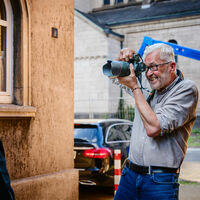 Achim Pohl Ein Mann fotografiert einen anderen Mann vor einer Hauswand mit Fenster. Eine Frau im Hintergrund assistiert ihm mit einem Reflexschirm.