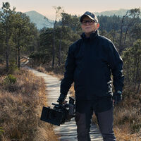 Ein Mann steht auf einem Holzbohlenpfad in einer offenen Waldlandschaft aus Kiefern und niedrigen Sträuchern und blickt in die Ferne. Er trägt Outdoor-Kleidung und eine Basecap und hält eine Filmkamera in der rechten Hand. Im Hintergrund sind Berge zu sehen.