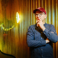 Ein Mann mit Brille und roter Kappe lehnt in nachdenklicher Pose an der Wand eines Kinosaals. Einen Arm hat er vor der Brust verschränkt, den Zeigefinger des anderen an den Mund geführt. Die Wand ist wellenförmig und in goldenes Licht getaucht. Ein opulenter Balkon ist zu sehen, zwei rote Stühle und blütenförmige, runde Wandlampen.