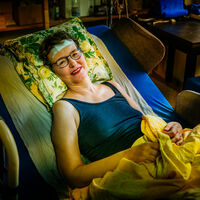 Claudia Wissmann Eine Frau liegt in einem Krankenbett, dessen Kopfteil hochgestellt ist. Sie hat ein Pflaster auf der Stirn und lächelt in die Kamera.