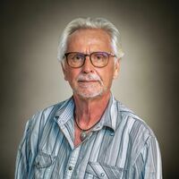 Portrait eines weißhaarigen Herrn mit Brille und Bart im hellblau gestreiften Hemd und Kette mit auffälligem grünen Stein. 