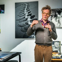 Matthias Kanter Mann mit gelber Brille, bunter Perlenkette und strubbeligem Haar redet lebhaft mit den Händen, die Augen geschlossen. An der Wand hängen verschiedene Schwarz-Weiß-Fotografien und farbige Gemälde. Auf einem Besprechungstisch liegen Aquarelle und eine Pfeife mit Tabak.