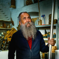 OMI Riesterer Älterer Mann mit grauem Rauschebart steht in einem Atelier und blickt visionär in die Ferne. Er hält eine große, runde Eisenstange in der Hand. Im Hintergrund sind viele gedrechselte oder quadratisch strukturierte Modelle aus Holz oder Metall in Regalen aufgereiht.